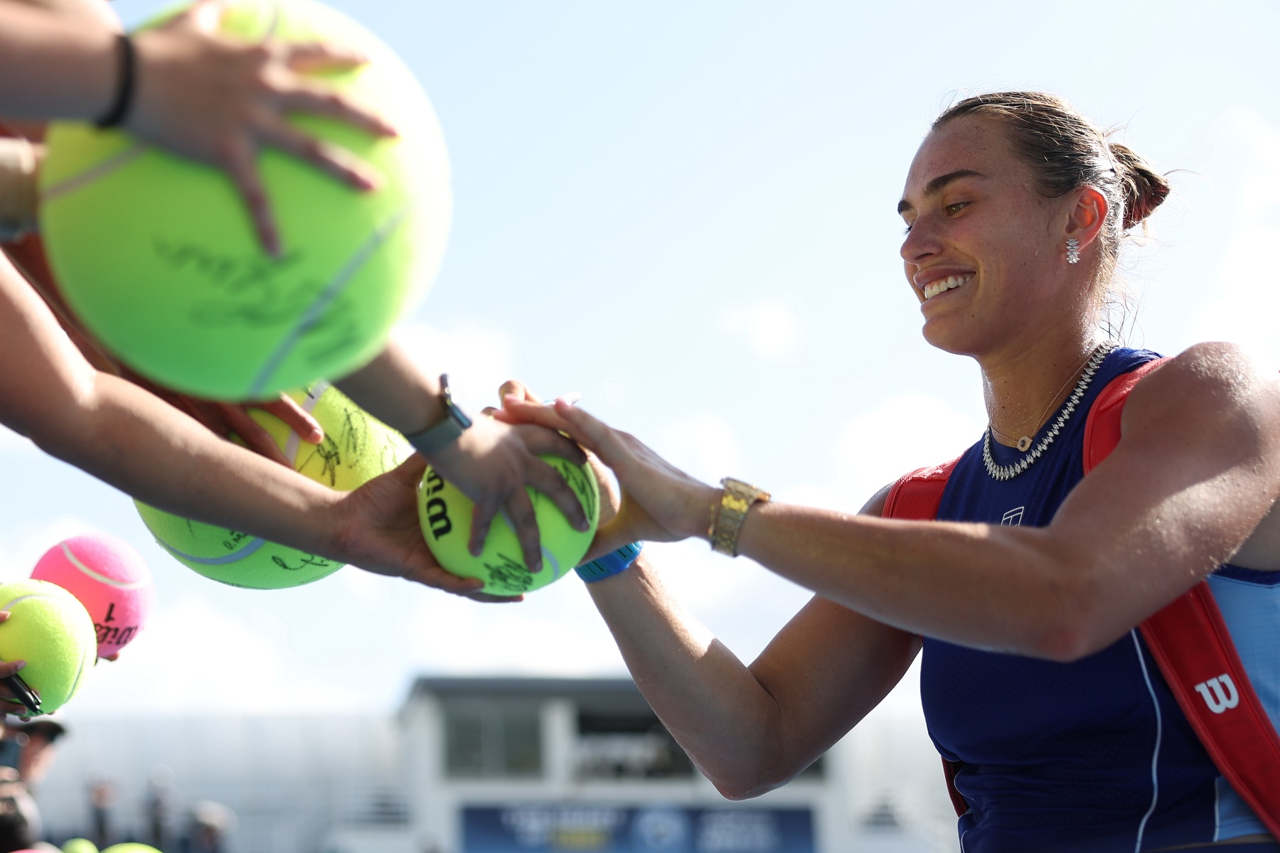 A player smiles as she signs oversized tennis balls held out by fans at the barrier, a kit bag over her shoulder.